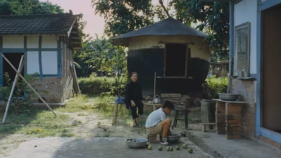 A young man crouches beside washbasins in a courtyard as an elderly woman sits nearby