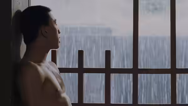 A shirtless young man looks out through a barred window at heavy rain