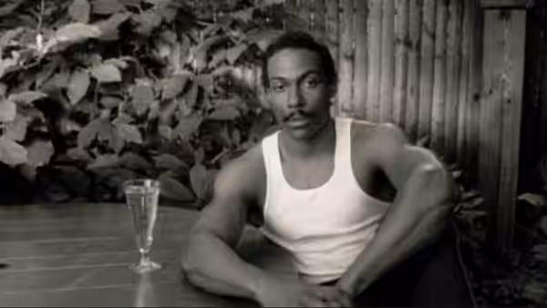 A mustached man in a tank top sits at a garden table beside a glass