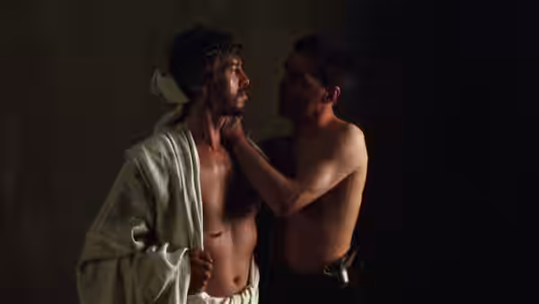 A shirtless man touches the face of a wounded man in a robe