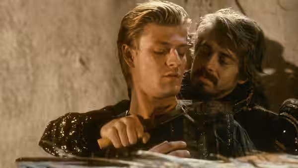 A dark-haired man holds a knife close behind a blond man