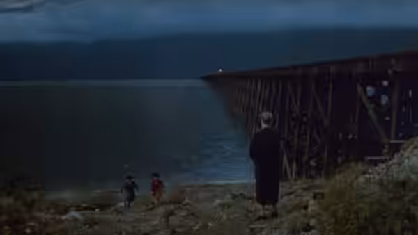 A woman watches two figures on the rocky lakeshore beside a long wooden trestle