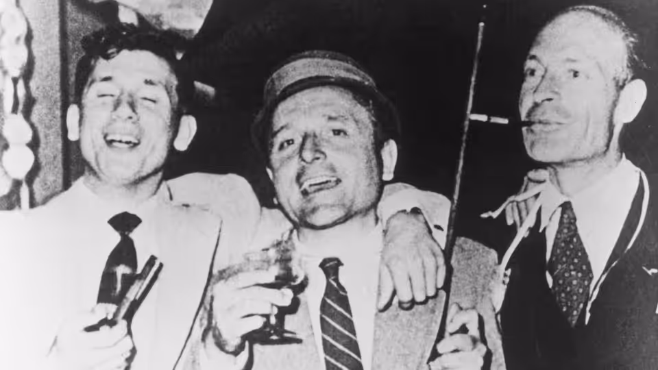 Three men in suits stand together, smiling and holding drinks at a gathering