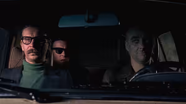 Three men sit inside a car, with one behind the wheel and two in the back