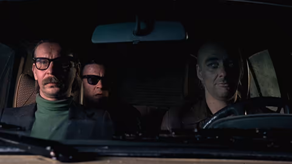 Three men sit inside a car, with one behind the wheel and two in the back