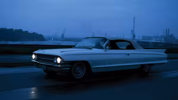 A white Cadillac drives along a road beside the harbour