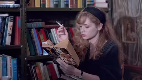 Audry (Adrienne Shelly) reads a book beside a library shelf, holding a cigarette
