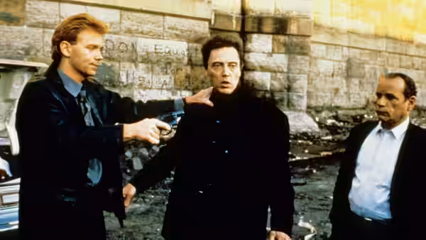 Frank White (Christopher Walken) stands between two men beside a stone wall