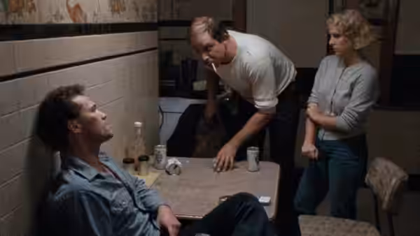 Henry (Michael Rooker) sits at a table as Otis leans over it and Becky stands nearby