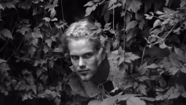 A blond man peers through dense foliage