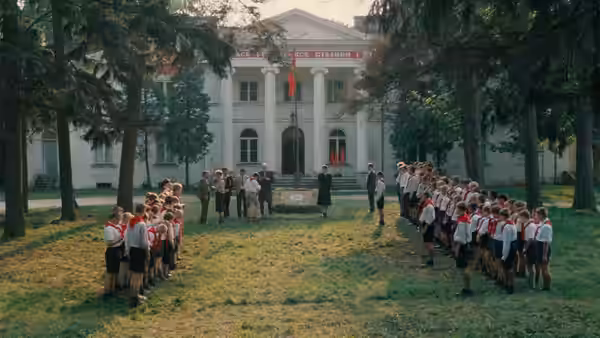 Children stand in uniformed lines outside a large building as adults gather between them