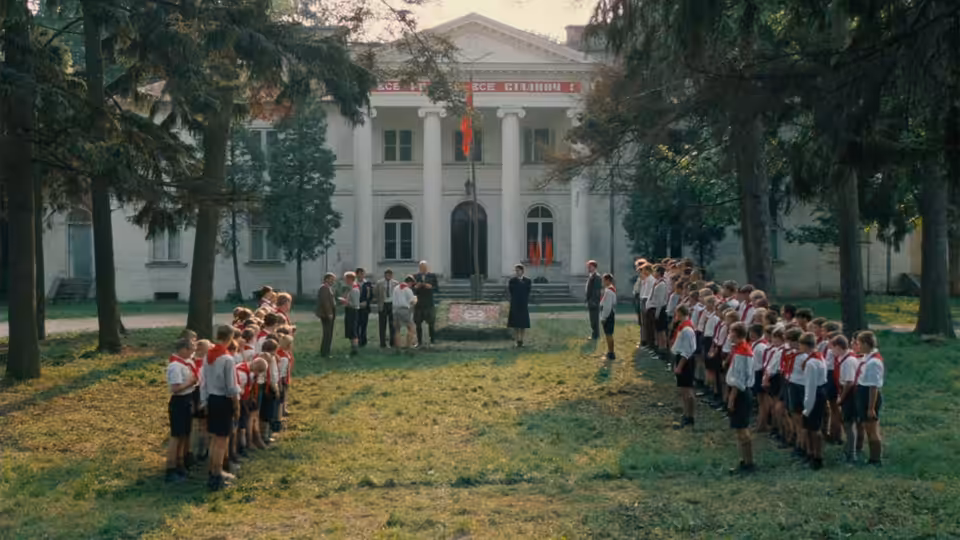 Children stand in uniformed lines outside a large building as adults gather between them