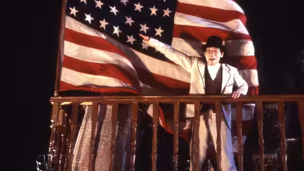 A man in a white suit gestures before a large American flag