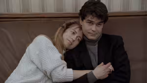 Félicie leans against a seated man, holding his arm indoors