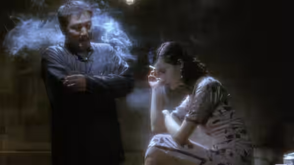 A man stands with folded arms as a seated woman smokes