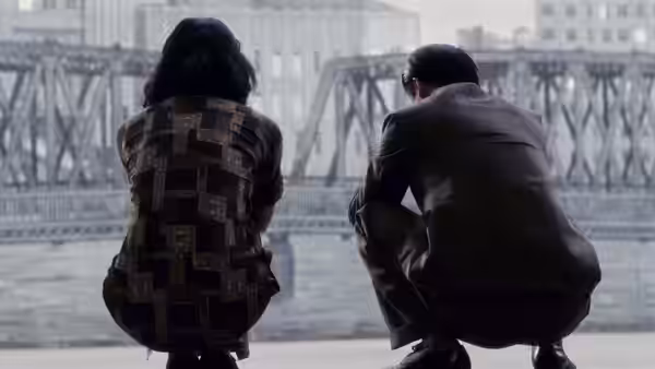 A woman and a man crouch beside a river, facing an iron bridge