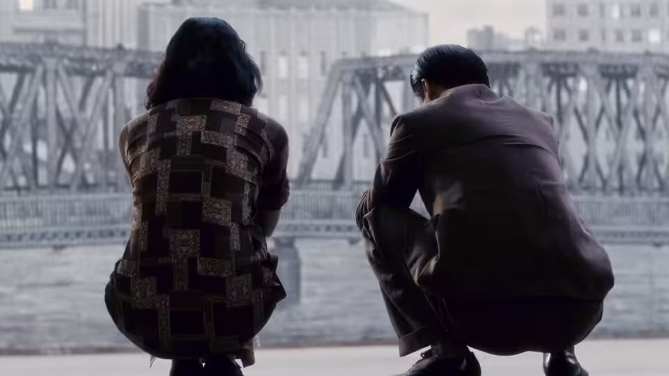 A woman and a man crouch beside a river, facing an iron bridge