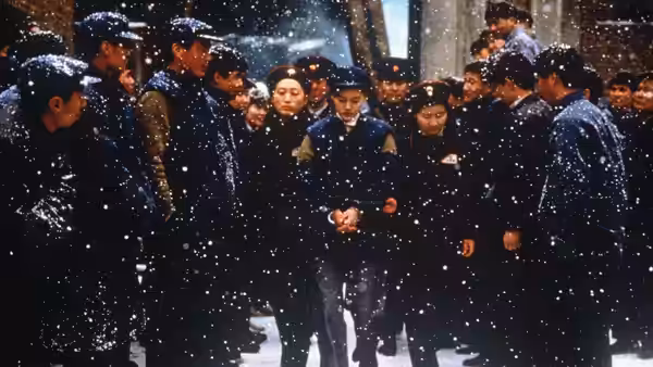 A woman stands among mourners in dark coats as snow falls in a courtyard