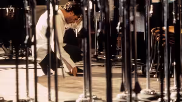 A man bends among microphone stands, reaching toward the floor in a recording studio