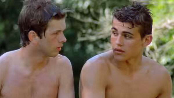Two shirtless young men sit together in a wooded area
