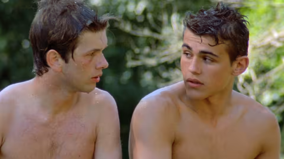 Two shirtless young men sit together in a wooded area