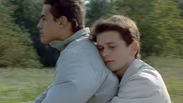 Two young men ride together outdoors, one resting his head against the other's back