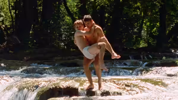 A young man carries another through a shallow, rocky stream