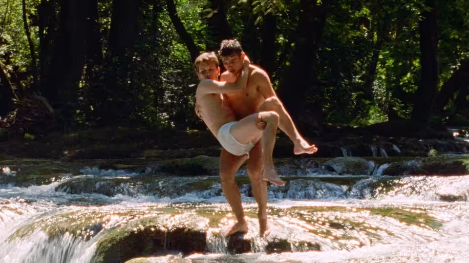 A young man carries another through a shallow, rocky stream