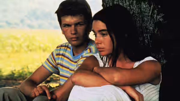 A young man sits beside a young woman beneath a tree as she hugs her knees