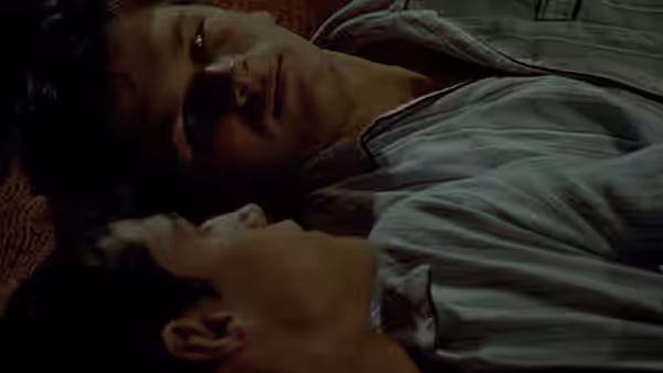 Two young men lie side by side indoors, gazing at each other