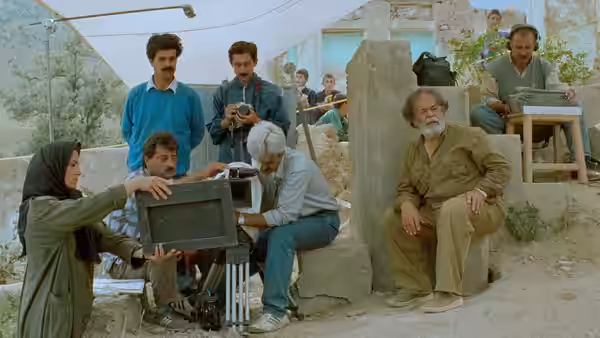 A film crew works beside an older man seated by a stone wall