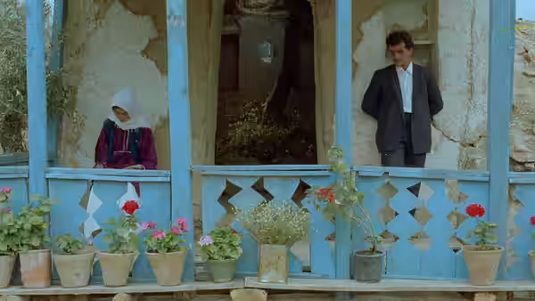A young woman and a suited man stand apart on a blue veranda