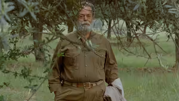 An older bearded man stands among olive branches holding a jacket