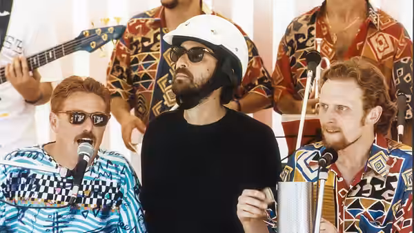 A helmeted man in sunglasses stands between two singers at microphones