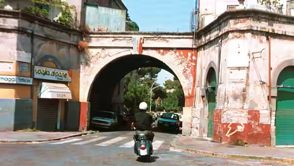 A scooter rider passes beneath an archway into a tree-lined street