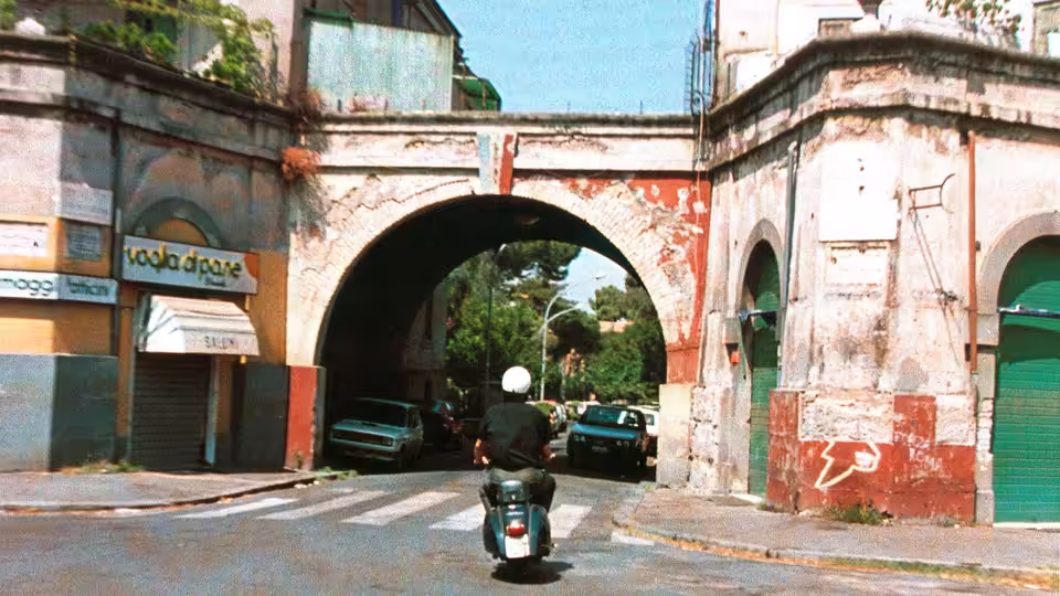 A scooter rider passes beneath an archway into a tree-lined street
