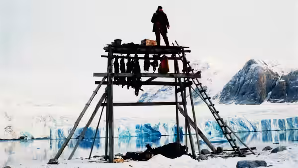A bundled man stands atop a wooden rack hung with meat beside the icy shore