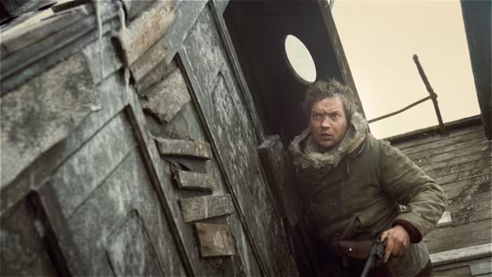 A bundled man crouches in the doorway of a weathered hut, holding a rifle