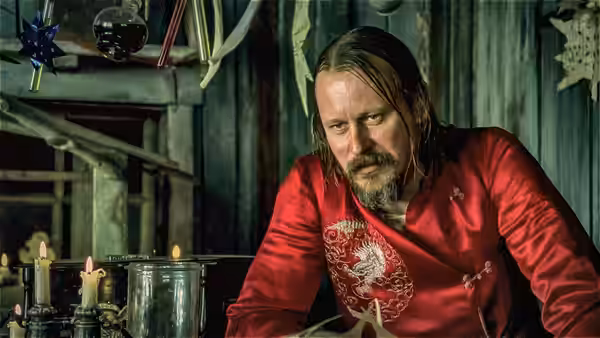 A long-haired, bearded man in a red shirt leans over a table inside a cabin