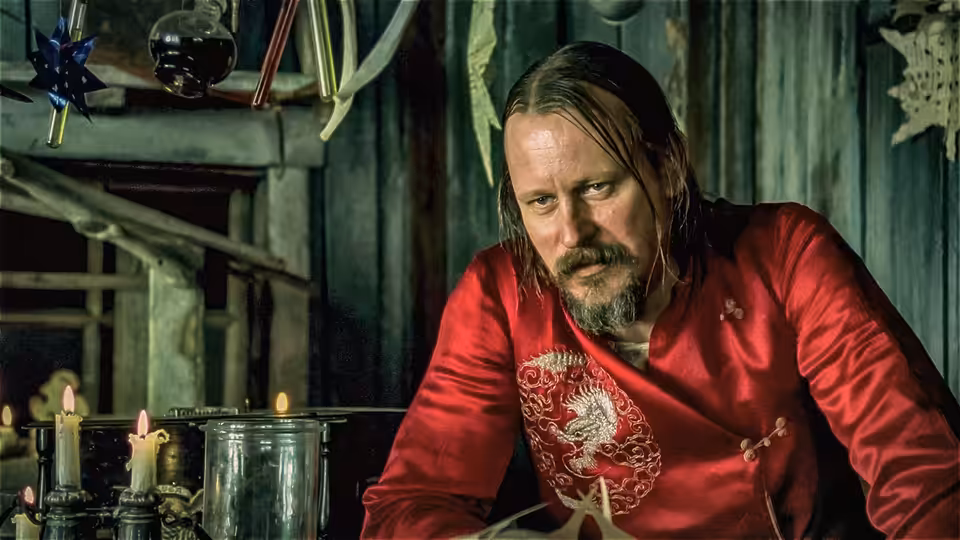 A long-haired, bearded man in a red shirt leans over a table inside a cabin
