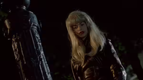 A blonde woman in a leather jacket stands beside a statue in the dark