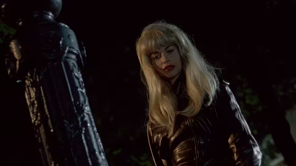 A blonde woman in a leather jacket stands beside a statue in the dark