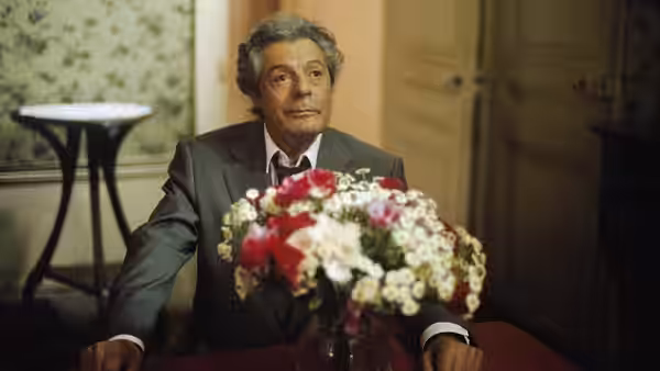 A grey-haired man in a suit sits behind a large bouquet of flowers