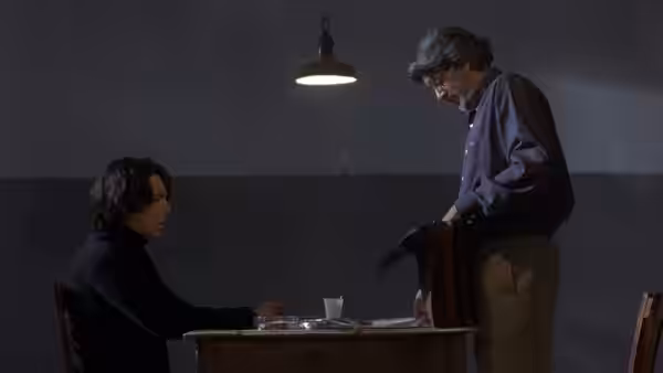 César sits at a table as an older man stands over him beneath a lamp