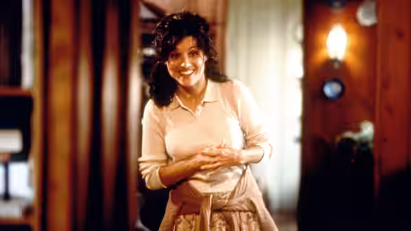 A smiling woman stands with her hands clasped in a warmly lit doorway