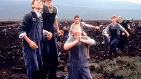 A boy kneels with clasped hands as children gather in a muddy field