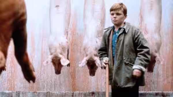 Francie Brady (Eamonn Owens) holds a stick beside hanging pig carcasses