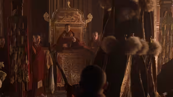 A young monk sits on an ornate throne as attendants gather inside the temple