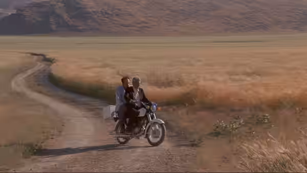 Two men ride a motorcycle along a dirt track through open fields