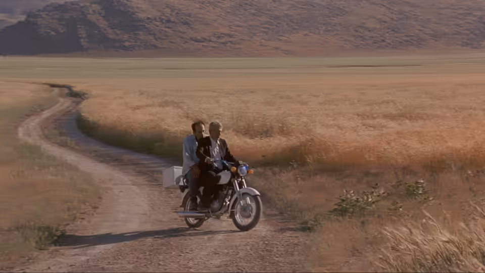 Two men ride a motorcycle along a dirt track through open fields